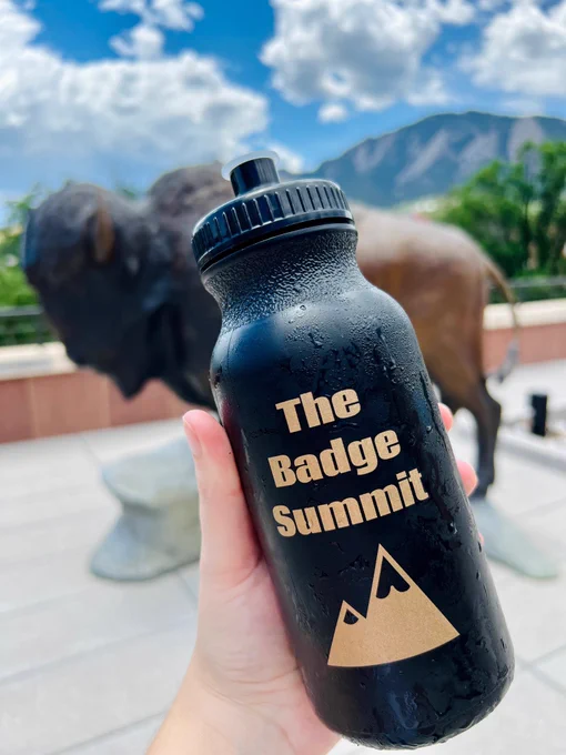Black and gold Badge Summit water bottle pretending to be the main attraction while clearly being overshadowed by the Buffalo statue and stunning mountains in the background that are the real allure to come to the conference in Boulder.