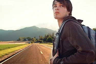 A young man looks back over his shoulder in the middle of a long, straight, two-lane highway leading towards trees and a mountain ahead.