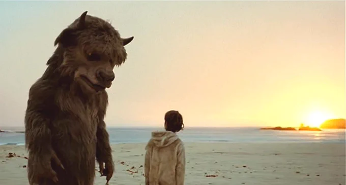 The Bull from Where The Wild Things Are stands next to Max. The Bull is a large monster who resembles a bull, but has human hands and is standing in two feet. He has dark grey and black fur. He and Max stand on the beach at sunset.