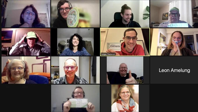 Screenshot eines Videoanrufs. 13 Personen lächeln in die Kameras. Einige halten Wheelmap Materialien hoch. 