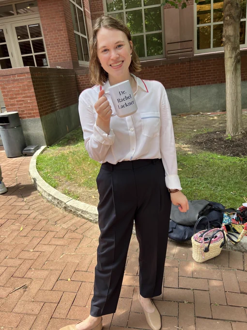 A woman in a white shirt and blue pants stands and smiles, holding a mug that says "Dr. Rachel Lackner" 