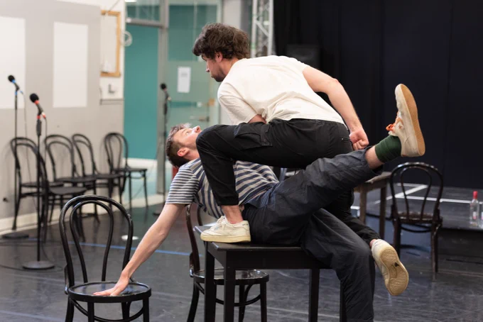 A rehearsal room, two men are on a table pretending to fight
