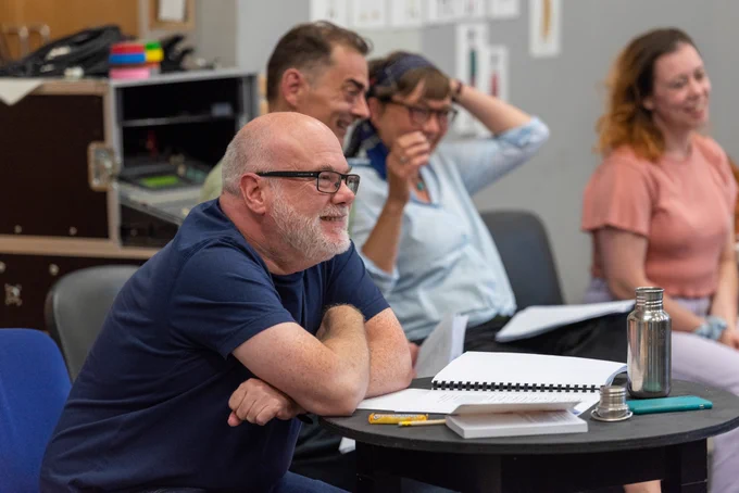 A group of people sitting at tables in a rehearsal room. 
