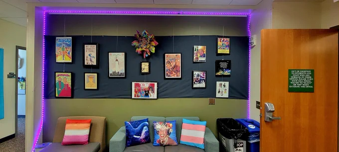 A photo of the Community Art Wall, which is framed by purple string lights, a dark gray cloth background, and holds approximately 13 pictures and 1 Passion Flower
