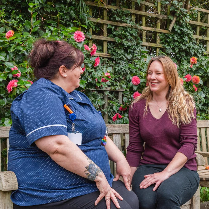 Nurse in blue uniform sat on a bench with a woman in a maroon jumper, both looking and smiling at each other.