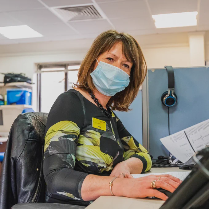 Woman wearing a patterned top and face mask, looking at the camera whilst sat at a desk, working on a computer.