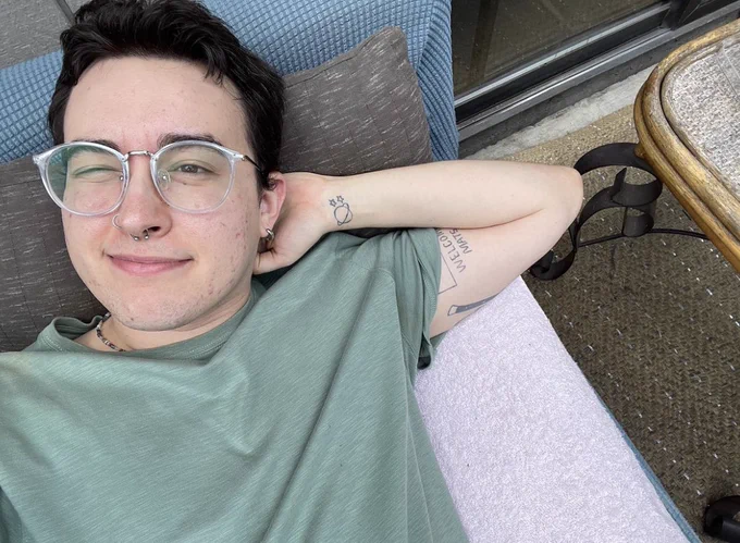 a chest up picture of Rowan. they are wearing an olive green shirt. his arm is bent behind his head and he’s squinting at the camera.