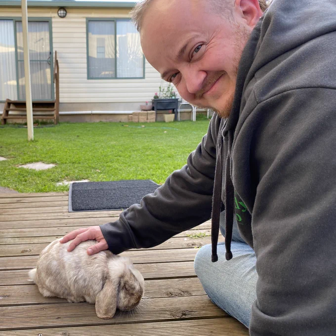 Person smiles at the camera while patting a wholemeal coloured fluffy rabbit with floppy ears