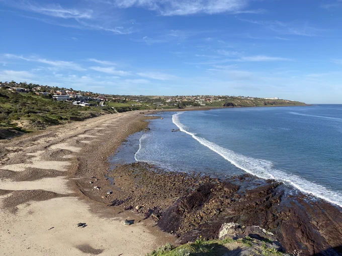 View from one end of a beach, with green land and houses to one side and a flat blue sea to the other. There is a single, long line of white foam cutting along the entire cove, close to shore.