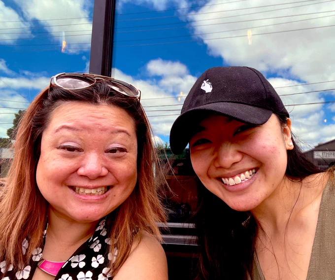 Marjorie & April smiling in front of a reflective mirror showing a sunny sky.