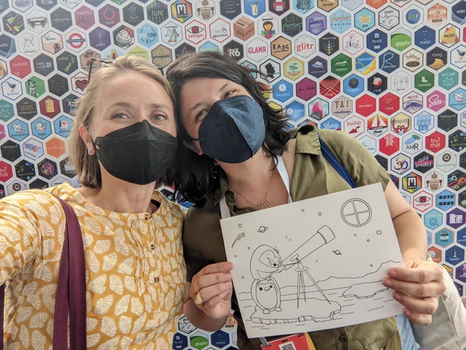 two women smiling with masks standing in front of a wall of hex shaped R packages stickers, and holding up a coloring page