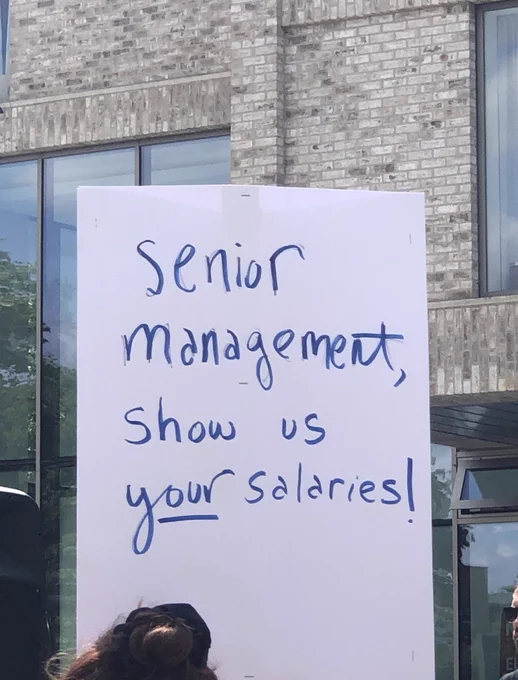 Protest sign that reads ‘senior management, show us your salaries!’. The word ‘your’ is underlined.