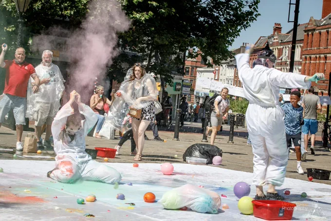 Image of two people wearing white overalls popping water balloons full of paint and chalk on a white sheet on the Preston flag market.