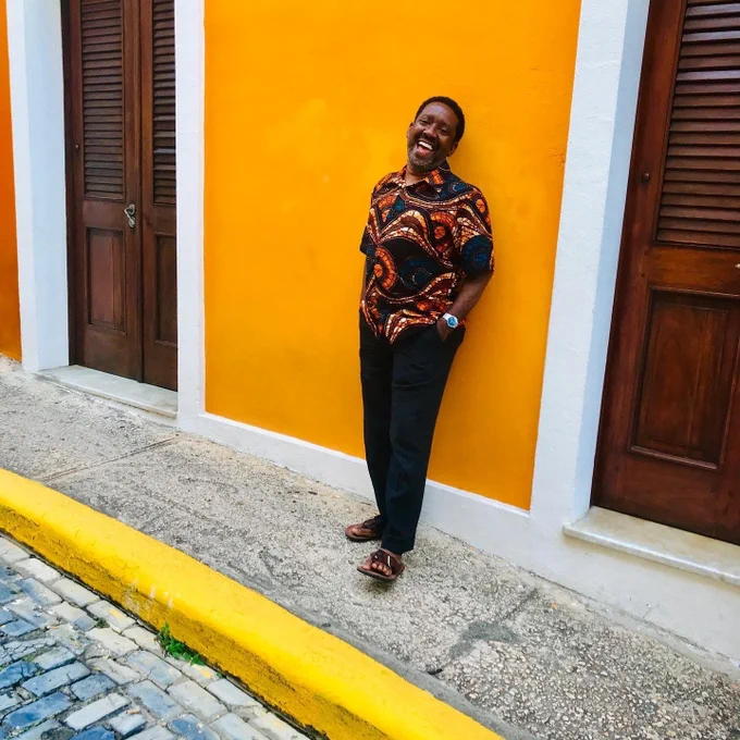 A photo of outgoing FFL president and CEO Flozell Daniels, Jr. He is standing on a sidewalk in front of a bright orange wall. He is smiling and looking directly into the camera. 