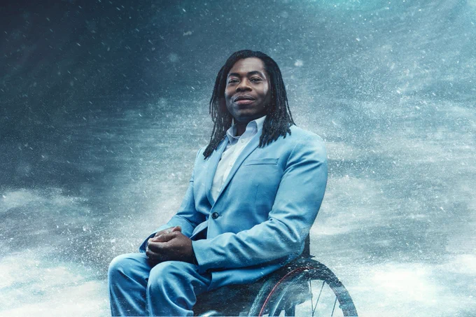 Black male presenter Ade Adepitan in a light blue suit sitting in his wheelchair