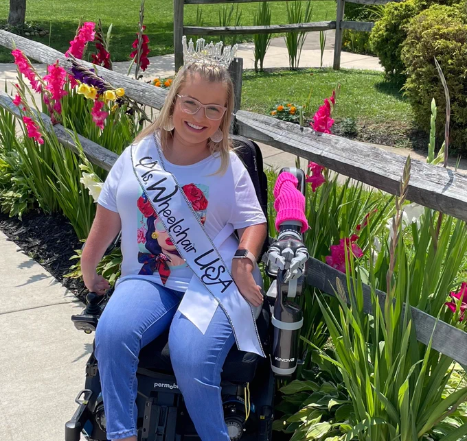Sara Gaver, Ms. Wheelchair USA, poses outside in her crown and sash in front of flowers.