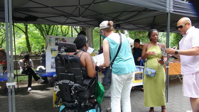 Two PIST volunteers described earlier have come around to the front of their table to be in conversation with a Black woman in a wheelchair on the left and a standing gentleman of color on the right.
