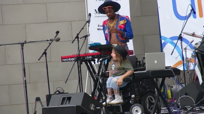 Two of the amazing performers on the Disability Unite stage who made the day so enjoyable.  Tall Black man in hat and colorful top is at the keyboards; small white woman in a wheelchair is at the microphone. Speakers, cords, and a laptop on a stand are nearby. 