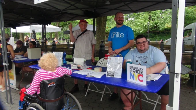 Three white men are at a table to promote Disability Pride NYC parade for October 2, 2022. Man at far left is making a thumbs-up gesture. Man in middle wears a turquoise Disability Unite volunteer T shirt. Man on right is seated and smiling. In front of the table is a woman in a wheelchair seen from the back she has curly blond hair. 