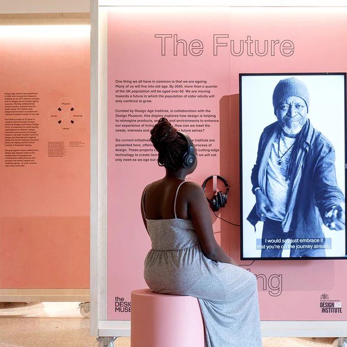 Woman with headphones on sitting on a pink stool,  watching a film playing as part of an exhibit.  