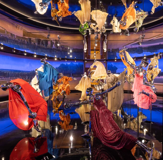 A view of a fashion exhibition at The Met featuring silver mannequins wearing long dresses and gowns as they engage in sword-fighting. A reflective ceiling and floor, as well as blue-tinted lighting, create a shimmery glow.