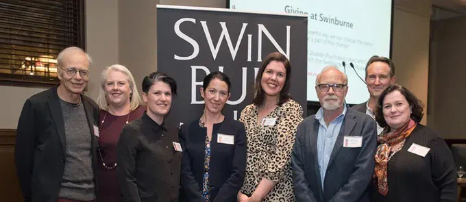 Eight people stand in a line in front of a Swinburne university banner. They are all wearing name tags and smart casual clothing. A powerpoint is visible in the background, with text reading; "Giving at Swinburne".
