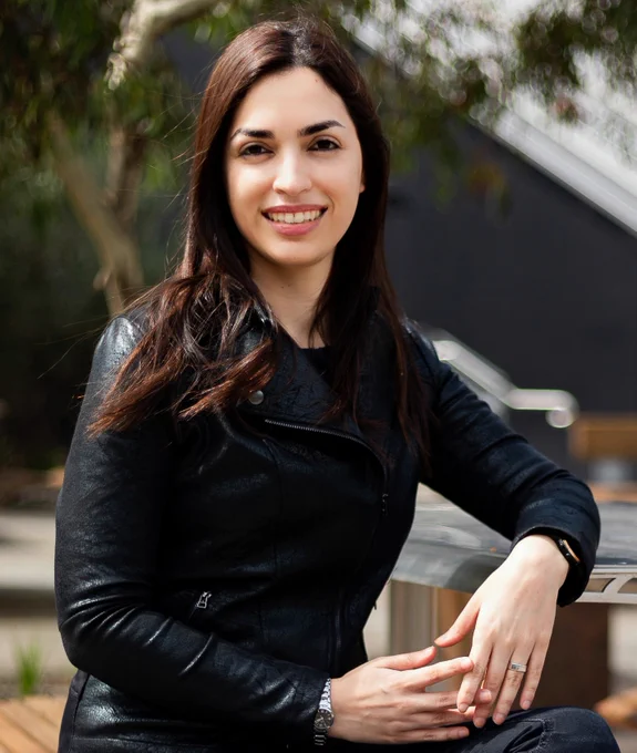 A woman sits outside in a back jacket with a tree in the background. 