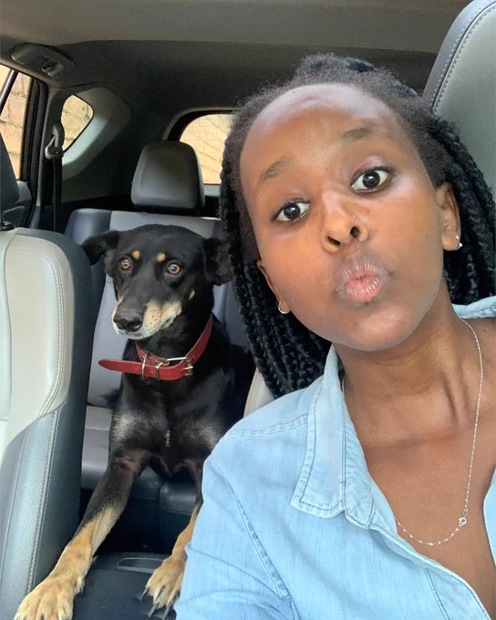 Angela sitting in the front seats of a car, wearing a blue shirt and posing with her dog.