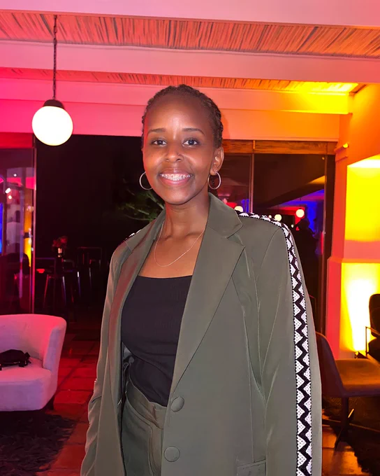 Angela wearing a green blazer with white and black on the arms, and smiling with a pink and orange lit up background.

