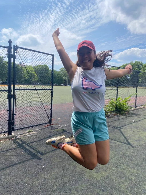 woman jumping in the air while wearing white dino tank top with pink dinosaur and the words "not extinct" in rainbow text. she looks very cool like she's having a great time.