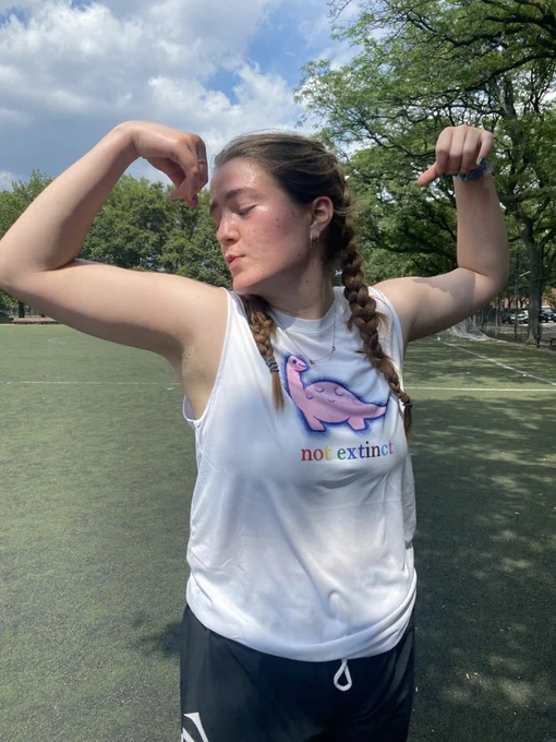 woman wearing white dino tank top with pink dinosaur and the words "not extinct" in rainbow text, she is miming kissing her arm muscles, she looks very strong and cool.