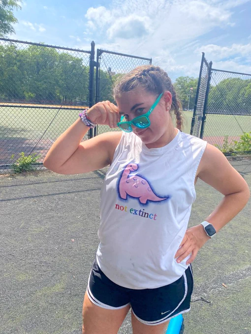 woman wearing white dino tank top with pink dinosaur and the words "not extinct" in rainbow text. she is holding sunglasses looking very cool.