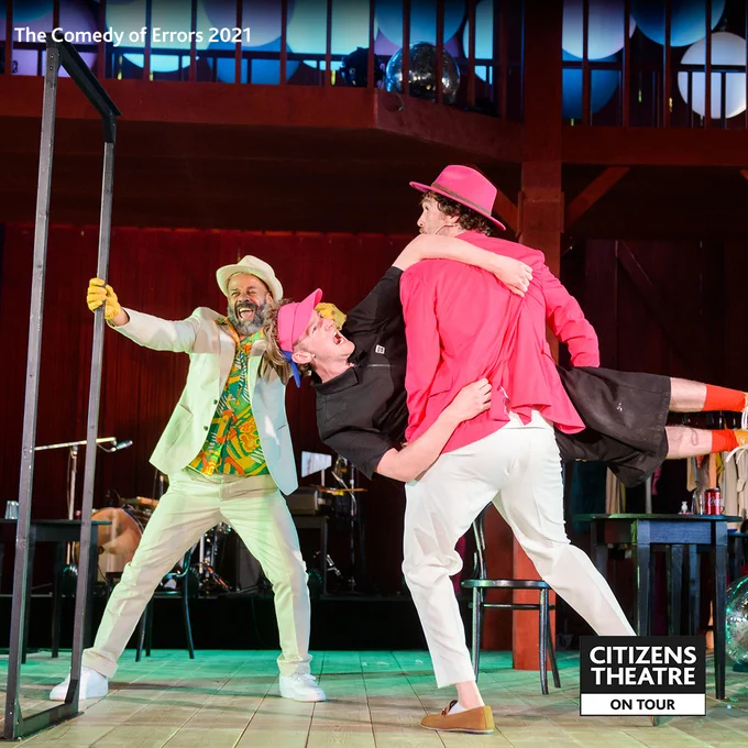 Three people are on a brightly lit stage. One wears a white suit with a white hat, one wears a pink suit with a pink hat and one wears a black t-shirt with a pink sun visor. The person in the black t shirt is being carried sideways by the person in the pink suit - the person in the white suit laughs.
