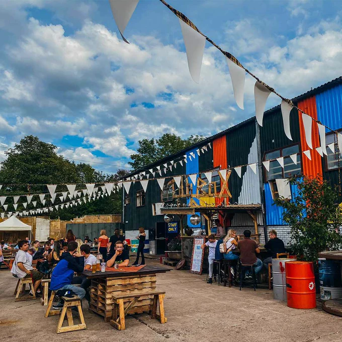Picture of the outside of the Kanteena venue, displaying picnic tables, white bunting, bright coloured walls and blue sky above. 