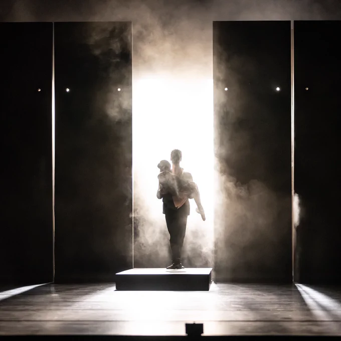 Two dancers emerge from the mist between two panels, one in the arms of the other.
