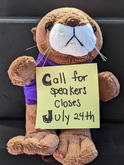 A stuffed toy of Mani the otter, holding a sing that says "Call for speakers closes July 24th"