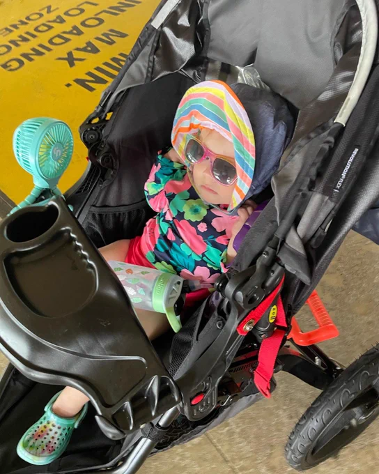 Toddler girl in a stroller, dressed in a colorful swimsuit, beach hat and sunglasses.