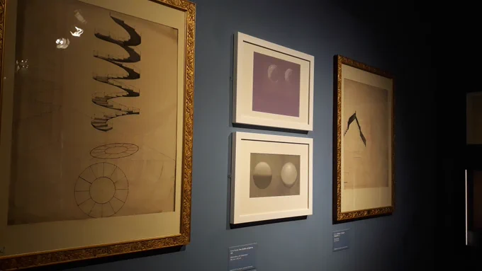 Two drawings of geometic shapes in gold frames; and two drawings of pairs of spheres in white frames. Hanging on a blue wall. 