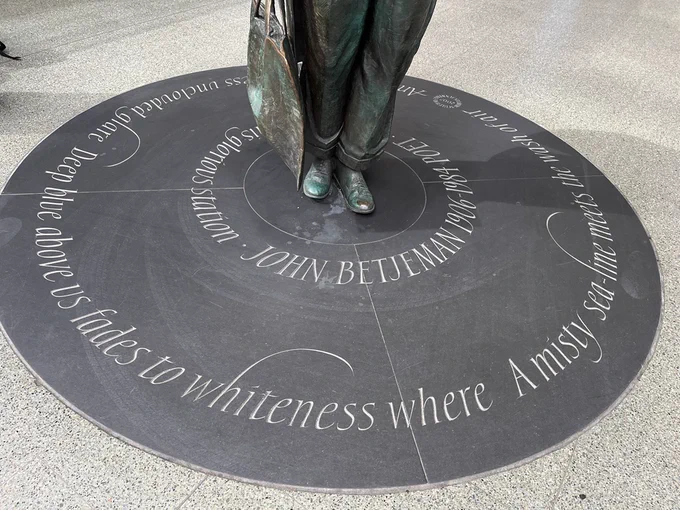 John Betjeman’s cast bronze feet stand at the centre of a slate circle with a quote from one of his poems circling round; the lettering is a seriffed italic with occasional flourishes