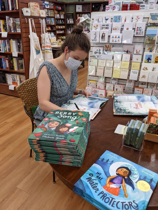 Michaela Goade sits at a round table, signing copies of her author-illustrator debut picture book, BERRY SONG. There is a stack of the book BERRY SONG in front of her. And just to the right of that are a few copies of WE ARE THE WATER PROTECTORS. There is a rack of cards behind the author, and bookcases filled with books and knick knacks in the background.
