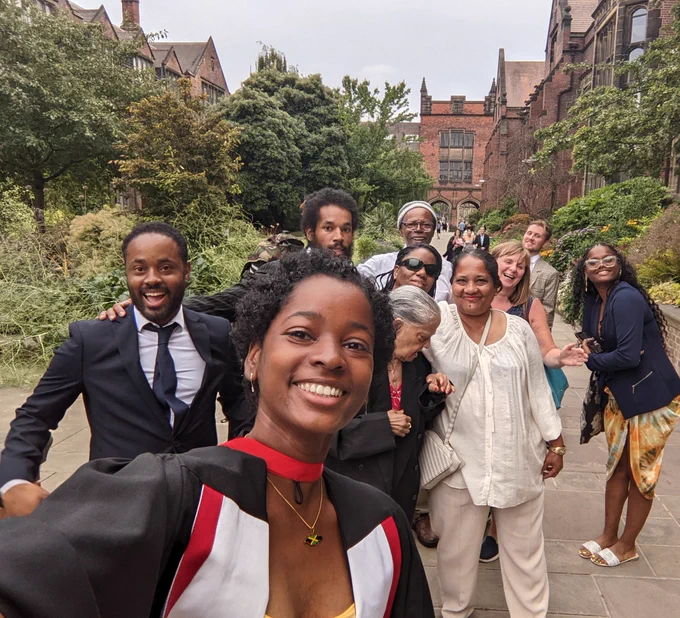 Group of 10 people smiling at the camera, in front of a university building and a lot of greenery.  