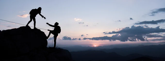 A stock photo of a person reaching out their hand to help another person to the top of a mountain. The sun is setting.