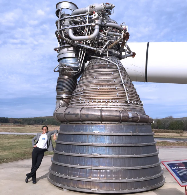 Dr. Bhavya Lal poses with a rocket engine. Wearing a grey professional jacket and dress pants Dr. Bhavya Lal leans on an F-1 rocket engine that towers three times her height.  The massive rocket engine is metallic grey and covered with pipes and bolts throughout. The circumference of the cone-shaped rocket is at least three times her height in length at its base and narrows towards the top. The rocket sits on a cement display and the background of the image is a wide-open field with a distant tree line and a clear blue sky visible. To the bottom right of the image is a blue placard with white text that reads "F-1 Engine". 