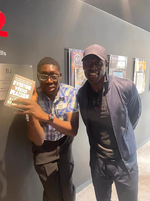 A young black teenager wearing glasses and a checked shirt holds a copy of a book to the camera. Next to him is an older black manwearing a baseball cap. He is the author of the book, Joshua Hutchinson.