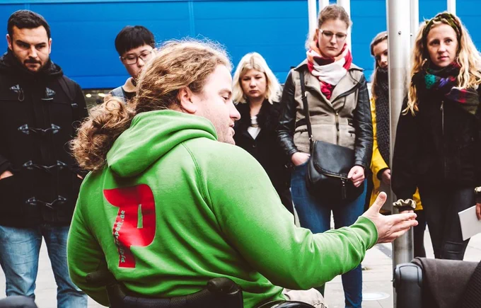 Ein junger Mann im Rollstuhl erklärt umstehenden Personen etwas. 