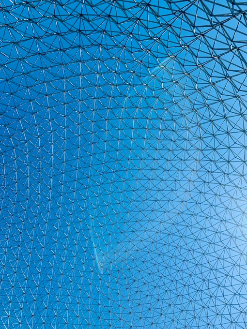 Photo from inside of the Biosphere, taken looking up. A transparent geodesic pattern fills the frame, with a blue sky behind it.