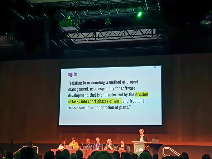Image shows speaker at lectern, with slides behind