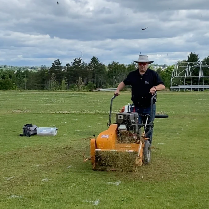 a man wearing a polo shirt and jeans pushes a verticutting machine- this machine resembles a snowblower