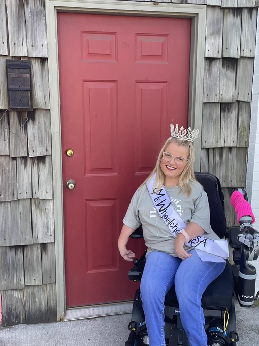 Sara Gaver smiles in front of a red door. She is wearing jeans and a pale green t-shirt with her Ms. Wheelchair USA crown and sash.