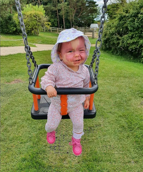Lily is sat in a swing, a lovely pink floral baby grow and hat. She's got bright pink shoes on.